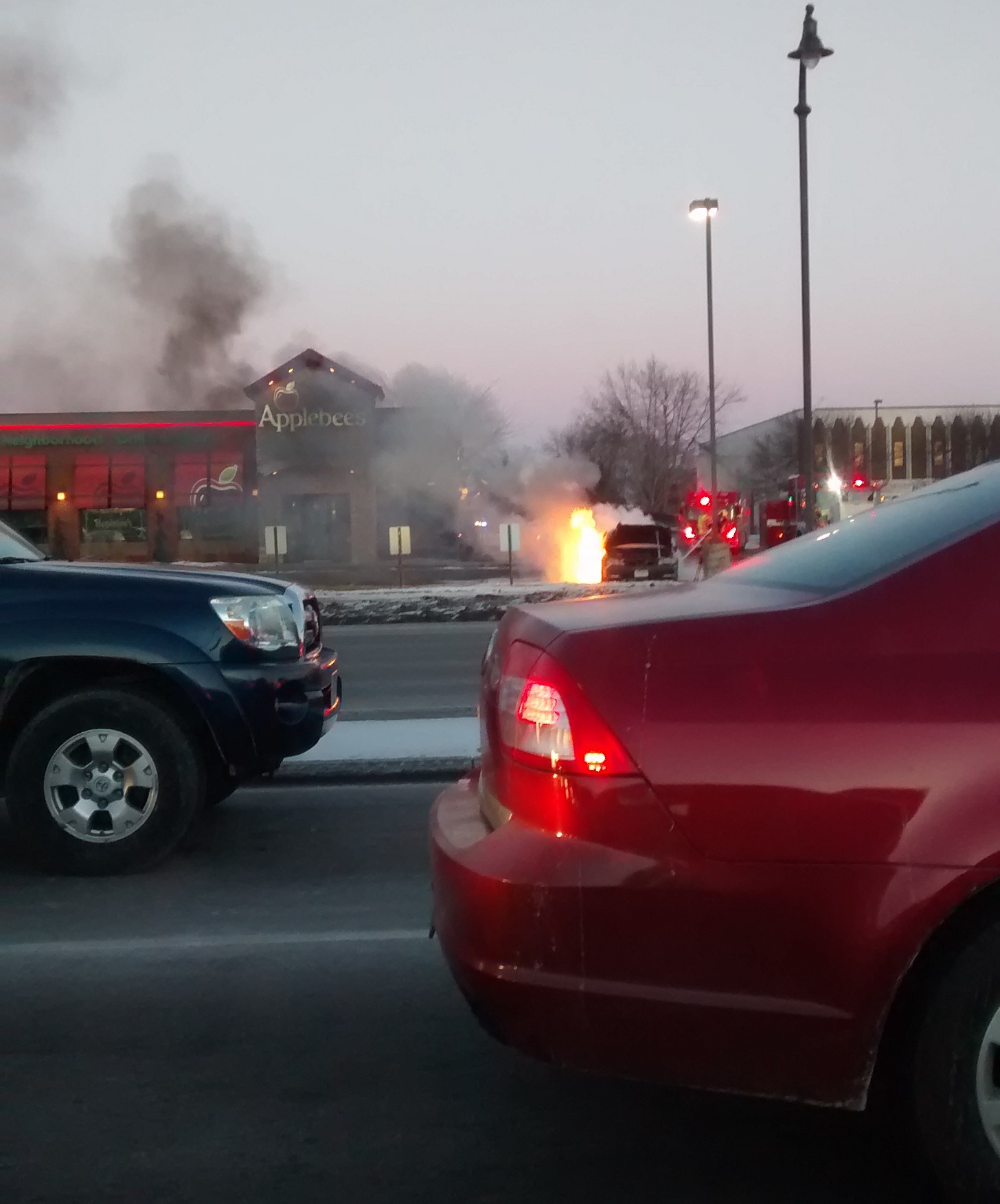 A car on fire in front of an Applebees.  Cars are patiently waiting in line to turn at a stoplight.  The scene has the feeling of the Wizard of Oz, 'Pay no attention to the man behind the curtain' 
